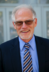 A smiling man with gray hair and glasses wears a dark suit, light blue shirt, and striped tie.