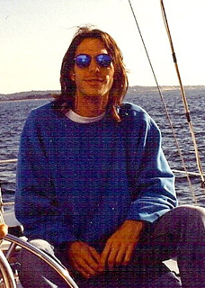 A man with long hair and blue sunglasses wearing a blue sweatshirt on a boat at sea.