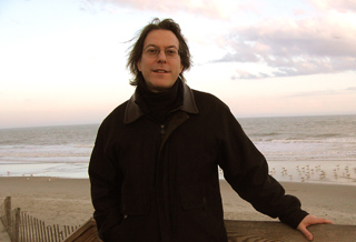 A man with glasses and dark hair wearing a black jacket stands on a beach against a sunset sky.