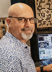A bald man with glasses and a grey goatee wearing a blue patterned shirt in a studio setting.