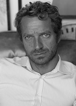 Black and white close-up portrait of a man with wavy hair and stubble wearing a striped button-down shirt.