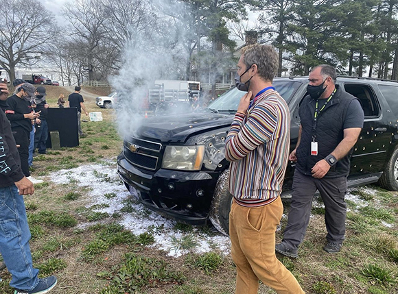 Smoke rises from the hood of a black SUV on a grassy field surrounded by several people.