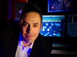 A man in a dark suit and white shirt smiling in a dimly lit music studio with monitors.