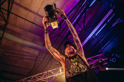 A tattooed person holds a large professional camera up toward the ceiling in a dimly lit venue.