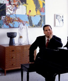 A man in a dark suit and red tie sits at a black piano in a room with large abstract paintings.