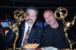 Two men smiling while holding gold Emmy awards at a table.