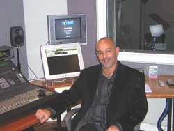 A man in a black blazer sits at a desk with a mixing console and computer monitors in a studio.