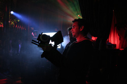 A man holds a professional video camera in a dark room with blue and red stage lighting.