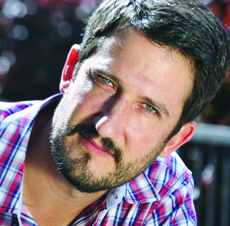Close-up portrait of a man with dark hair, a beard, and a pink and blue plaid shirt looking at the camera.