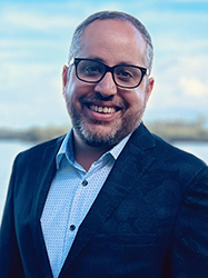 A smiling man with short hair and a beard wearing a dark blazer and light blue button-down shirt.
