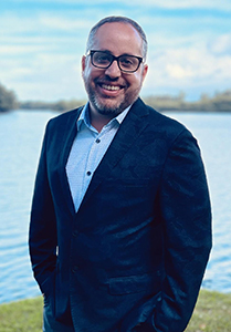 A smiling man with glasses and a beard wearing a dark blazer and light blue shirt, standing by a lake.