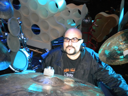 A man with glasses and a goatee wearing headphones behind a drum set.