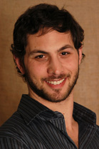 A smiling man with dark wavy hair and a beard wearing a dark striped shirt.