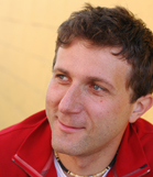 A man with short brown hair and blue eyes wearing a red jacket, looking off-camera.