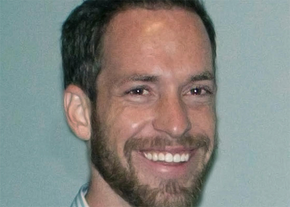 A smiling man with short dark hair and a brown beard looking toward the camera.