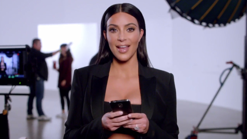 A woman with long dark hair wearing a black blazer holds a smartphone in a bright studio.