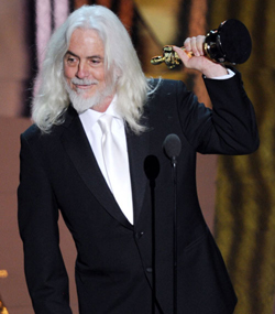 A man with long white hair and a beard in a black suit holds up a gold trophy.
