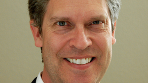 A close-up headshot of a middle-aged man with short brown hair and a wide smile.