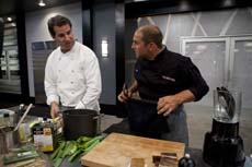 Two male chefs in white and black uniforms talk over kitchen counters with cooking tools.