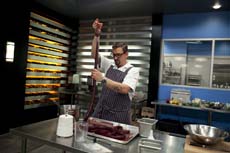 A man in a white shirt and striped apron pulls a long piece of dark meat upward in a professional kitchen.