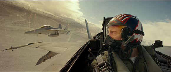 A pilot in a cockpit wearing a helmet and oxygen mask looks at a fighter jet flying over mountains.