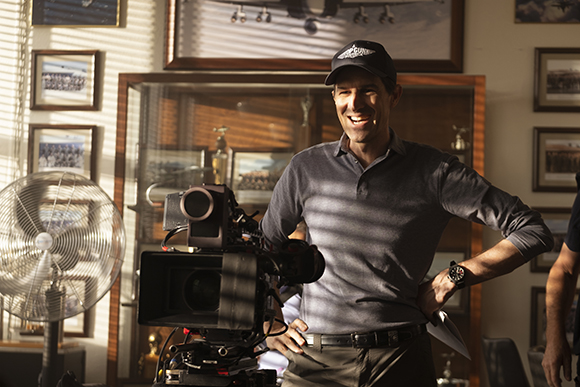 A smiling man in a grey polo shirt stands behind a large professional film camera in a sunlit room.