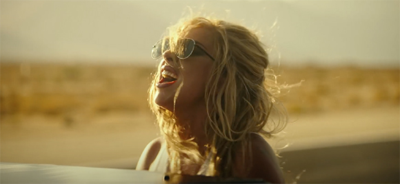A woman with blonde hair and sunglasses laughs while looking up toward the sun in a desert setting.