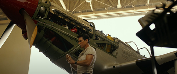 A man in a tan t-shirt examines the exposed engine and internal components of a large propeller plane.