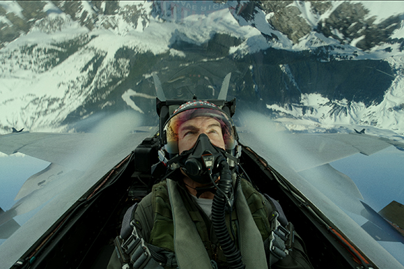 A pilot wearing a helmet and oxygen mask inside a fighter jet cockpit flying over snowy mountains.
