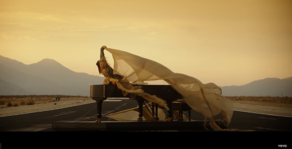 A person sits on a black grand piano in a desert, holding up a long, flowing white veil against a sunset sky.