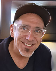A smiling man with glasses, a goatee, and a dark baseball cap looking toward the camera.