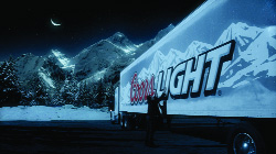 A Coors Light semi-truck parked at night against a backdrop of snowy mountains under a crescent moon.