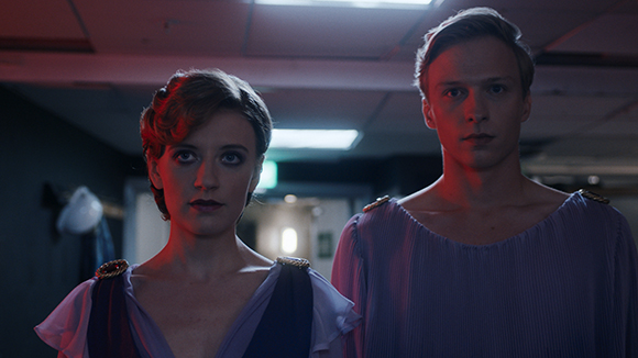 A woman and a man in purple classical garments looking forward in a dimly lit room.