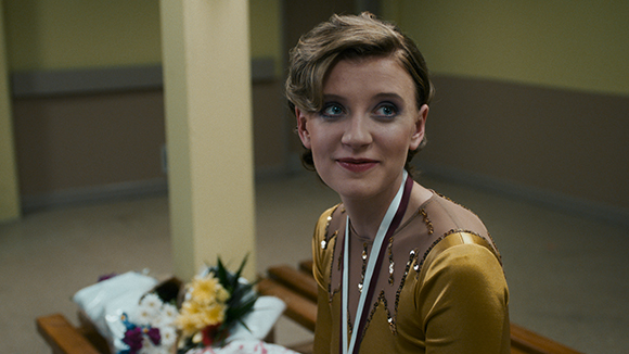 A young woman with short wavy hair wearing a gold figure skating outfit and a medal.