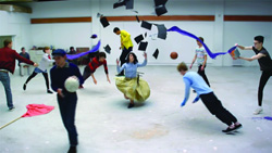 Performers move dynamically in a large room with graduation caps, balls, and blue fabric.