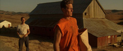 A man in an orange shirt stands in the foreground with another man and a large barn in the background.