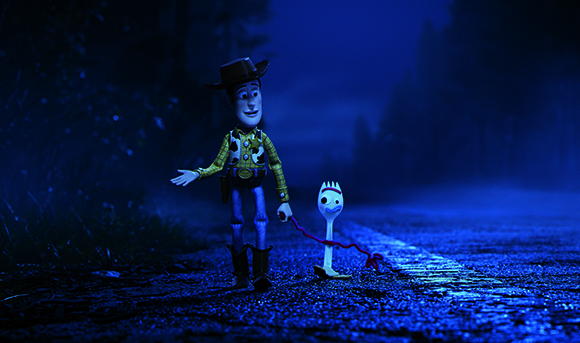 A cowboy toy and a small white spork toy stand on a dark, blue-lit road at night.