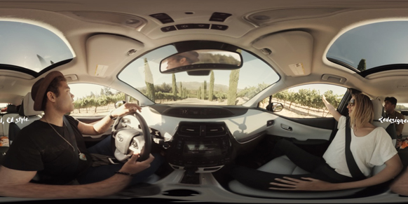 A man drives a car while a woman sits in the passenger seat, driving through a sunny vineyard.