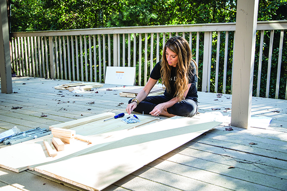 A woman sits on a wooden deck, using a screwdriver to work on light-colored wooden planks.