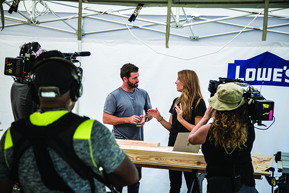 A film crew uses cameras to record a man and a woman standing in front of a white backdrop with a Lowe's logo.