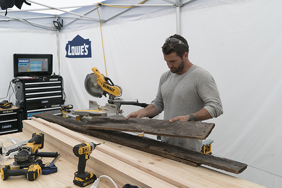 A man uses a miter saw to cut a piece of weathered wood on a workbench.