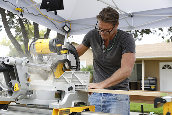 A man in a grey shirt uses a yellow and silver miter saw to cut a piece of lumber under a white canopy.
