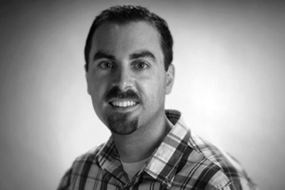 Black and white headshot of a man with dark hair, a mustache, and a small goatee, wearing a plaid shirt.