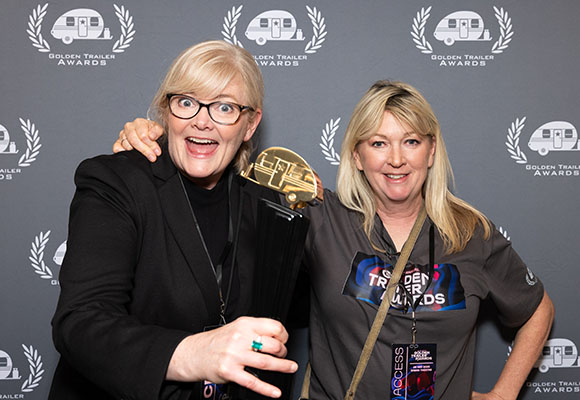 Two women smiling and posing together in front of a Golden Trailer Awards backdrop.