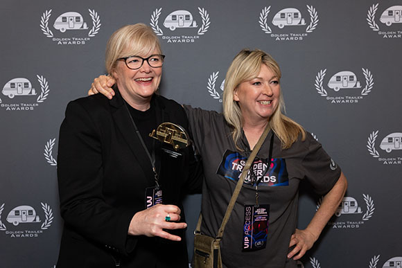 Two women smiling and posing together in front of a Golden Trailer Awards backdrop.