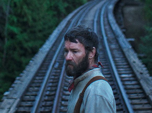 A man with a beard looks off to the side beside curved railroad tracks in a forest.