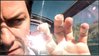 A man with a serious expression reaches his hand toward the camera lens under bright sunlight.