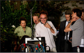 Several men stand outdoors near a camera rig and film equipment amidst green foliage.