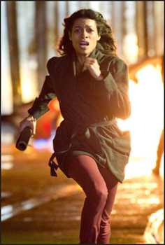 A woman with dark hair runs through a forest at night with a bright light behind her.