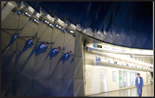 A man in a suit walks past a large, geometric mirrored wall reflecting multiple versions of himself.